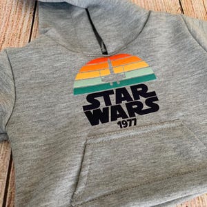 May include: Gray hooded sweatshirt featuring a colorful graphic of a sunset with a spaceship silhouette and the words "STAR WARS 1977" in black. The sweatshirt has a front pocket and a hood.