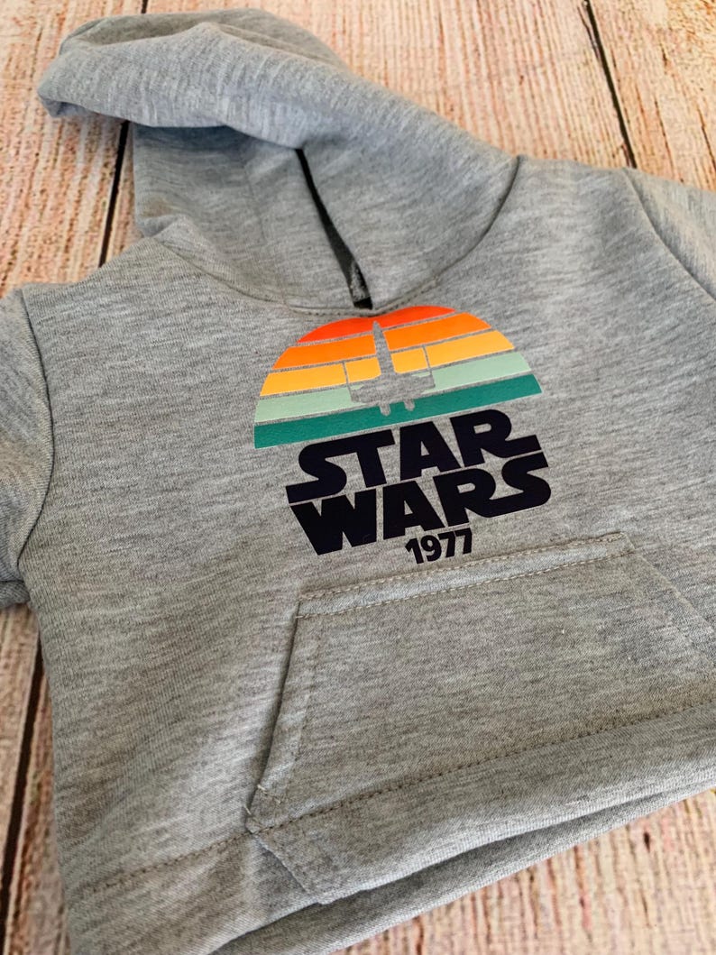May include: Gray hooded sweatshirt featuring a colorful graphic of a sunset with a spaceship silhouette and the words "STAR WARS 1977" in black. The sweatshirt has a front pocket and a hood.