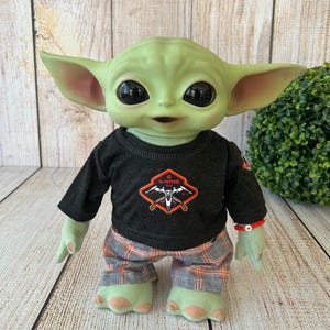 May include: A plush doll of a green-skinned alien character with large black eyes and pointed ears. The doll wears a black long-sleeved shirt with an X-Wing logo and plaid pants. A red beaded bracelet adorns its wrist.