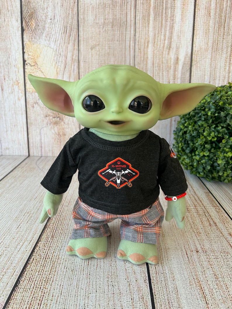 May include: A plush doll of a green-skinned alien character with large black eyes and pointed ears. The doll wears a black long-sleeved shirt with an X-Wing logo and plaid pants. A red beaded bracelet adorns its wrist.
