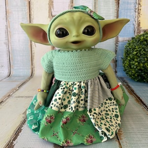 Puede incluir: Una muñeca verde con grandes ojos negros, vestida con un top de ganchillo verde menta y una falda patchwork con telas florales y estampadas. La muñeca tiene una diadema a juego con un lazo y pequeñas pulseras.