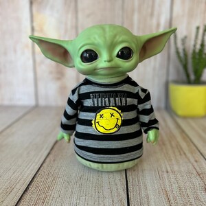 Peut inclure: Une figurine verte avec de grands yeux noirs et de grandes oreilles, portant un t-shirt ray&eacute; gris et noir avec le mot "NIRVANA" et un smiley jaune. La figurine est un personnage populaire d'une franchise de science-fiction.