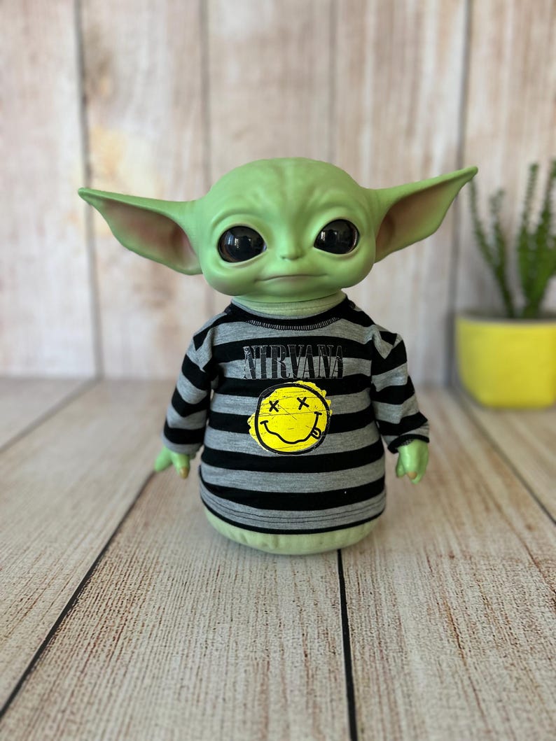 Peut inclure: Une figurine verte avec de grands yeux noirs et de grandes oreilles, portant un t-shirt ray&eacute; gris et noir avec le mot "NIRVANA" et un smiley jaune. La figurine est un personnage populaire d'une franchise de science-fiction.