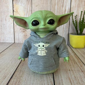 May include: A green figure with large black eyes and pointed ears, wearing a gray hoodie with the text "This is the way" and a graphic of the figure. The figure is on a wooden surface.