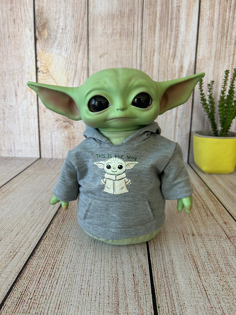 May include: A green figure with large black eyes and pointed ears, wearing a gray hoodie with the text "This is the way" and a graphic of the figure. The figure is on a wooden surface.