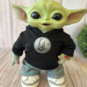 Op de afbeelding: Een groene wezenpop met grote donkere ogen en puntige oren, gekleed in een zwarte hoodie met een rond zilveren embleem met een schedelontwerp, een grijs en groen geruite broek en witte sneakers met grijze accenten op de neuzen.