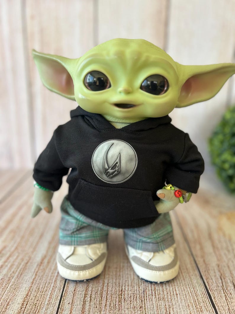 Op de afbeelding: Een groene wezenpop met grote donkere ogen en puntige oren, gekleed in een zwarte hoodie met een rond zilveren embleem met een schedelontwerp, een grijs en groen geruite broek en witte sneakers met grijze accenten op de neuzen.