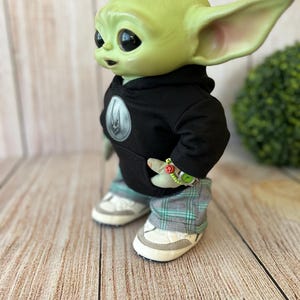 Op de afbeelding: Een groen wezenfiguurtje met grote donkere ogen en puntige oren, gekleed in een zwarte hoodie met een rond embleem, een grijs en groen geruite broek en witte sneakers. Een klein kralenarmbandje met een smiley-bedeltje zit om de pols.