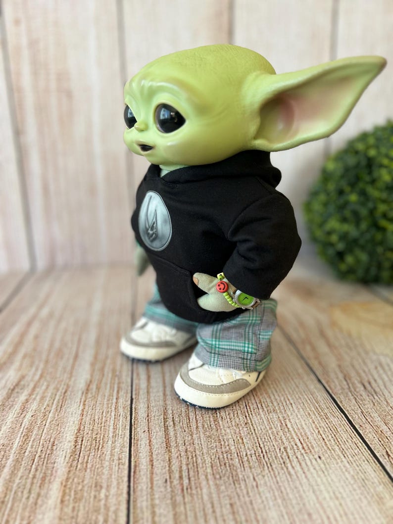 Op de afbeelding: Een groen wezenfiguurtje met grote donkere ogen en puntige oren, gekleed in een zwarte hoodie met een rond embleem, een grijs en groen geruite broek en witte sneakers. Een klein kralenarmbandje met een smiley-bedeltje zit om de pols.