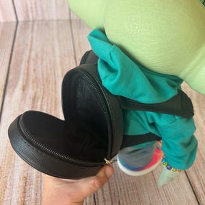 May include: A green-skinned character figure with large ears, wearing a teal hooded sweatshirt and a light blue hair bow. The figure carries a small, round, black zippered backpack with a textured surface and black straps on its back.
