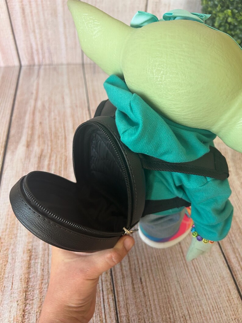 May include: A green-skinned character figure with large ears, wearing a teal hooded sweatshirt and a light blue hair bow. The figure carries a small, round, black zippered backpack with a textured surface and black straps on its back.