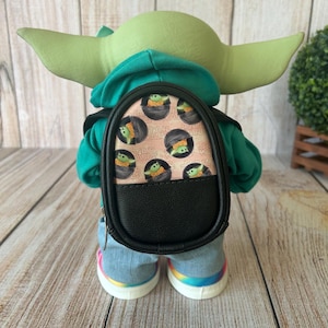 May include: A green-skinned figure with large ears wearing a teal hoodie, light blue denim pants, and white sneakers with rainbow soles. The figure carries a small black backpack featuring a repeating pattern of a green creature in a pod.