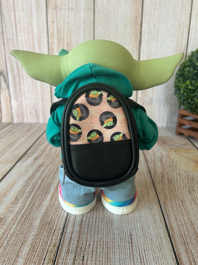 May include: A green-skinned figure with large ears wearing a teal hoodie, light blue denim pants, and white sneakers with rainbow soles. The figure carries a small black backpack featuring a repeating pattern of a green creature in a pod.