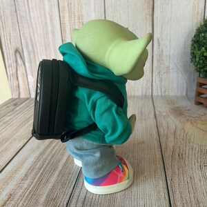 May include: A green-skinned creature figure wearing a bright teal hooded sweatshirt, grey pants, and sneakers with a rainbow-colored pattern. The figure carries a small black backpack with adjustable straps on its back, viewed from a side profile.