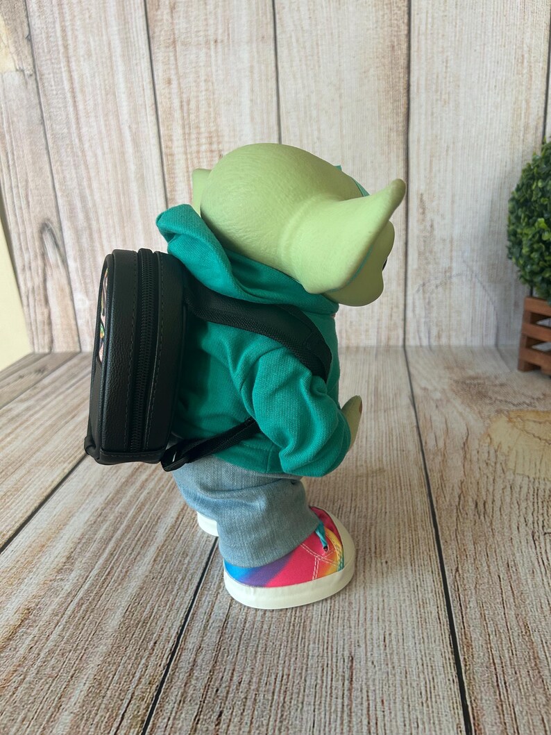 May include: A green-skinned creature figure wearing a bright teal hooded sweatshirt, grey pants, and sneakers with a rainbow-colored pattern. The figure carries a small black backpack with adjustable straps on its back, viewed from a side profile.