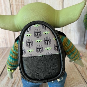 May include: A small backpack featuring a gray pattern of green alien characters and silver helmets, with a solid black bottom section. The backpack is worn by a green-skinned figure with large ears, wearing a striped sweater and blue jeans.