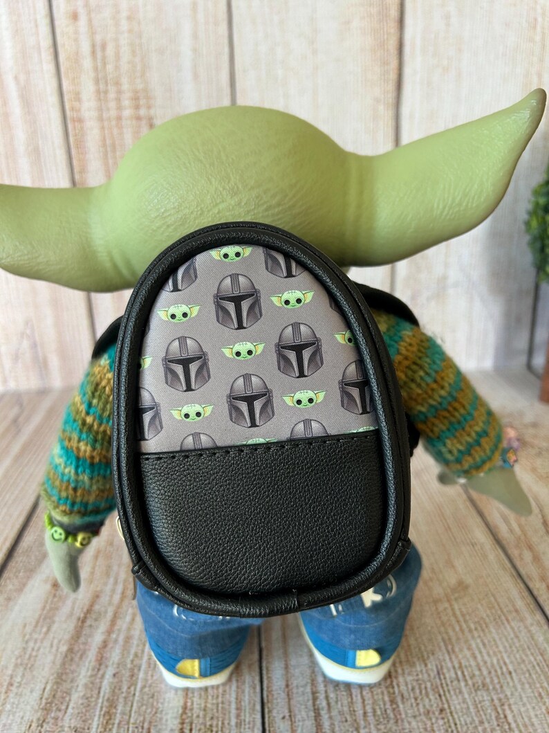 May include: A small backpack featuring a gray pattern of green alien characters and silver helmets, with a solid black bottom section. The backpack is worn by a green-skinned figure with large ears, wearing a striped sweater and blue jeans.
