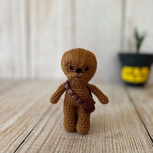 Könnte beinhalten: Eine braune gehäkelte Chewbacca-Figur mit einem schwarzen Bandolero. Die Figur steht auf einer Holzoberfläche.