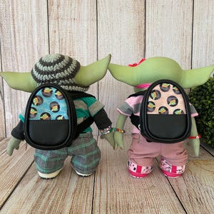 May include: Two green alien dolls standing side by side, each wearing a small black backpack. The left doll wears a gray and black striped crochet hat, a teal and gray shirt, and plaid pants. The right doll wears a pink shirt and pants.