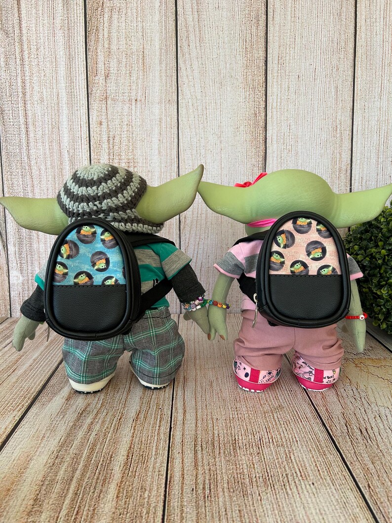 May include: Two green alien dolls standing side by side, each wearing a small black backpack. The left doll wears a gray and black striped crochet hat, a teal and gray shirt, and plaid pants. The right doll wears a pink shirt and pants.