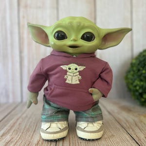 Op de afbeelding: Een groene wezenpop met grote zwarte ogen en puntige oren, gekleed in een mauve hoodie met een afbeelding van hetzelfde wezen en de tekst 'This is the way', gecombineerd met een groen-zwarte geruite broek en witte sneakers met grijze accenten.