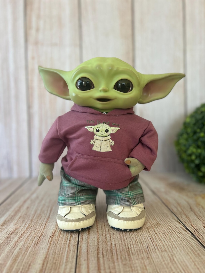 Op de afbeelding: Een groene wezenpop met grote zwarte ogen en puntige oren, gekleed in een mauve hoodie met een afbeelding van hetzelfde wezen en de tekst 'This is the way', gecombineerd met een groen-zwarte geruite broek en witte sneakers met grijze accenten.