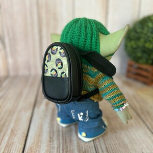May include: A small doll with green skin wearing a green crocheted hat, a striped knit sweater, and blue jeans. The doll carries a black backpack with a patterned front panel featuring small green creatures in black pods and black trim.