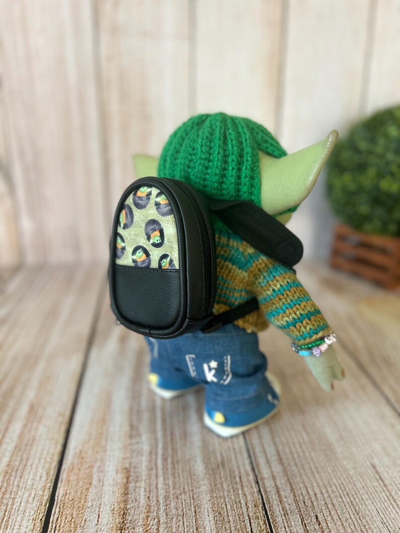 May include: A small doll with green skin wearing a green crocheted hat, a striped knit sweater, and blue jeans. The doll carries a black backpack with a patterned front panel featuring small green creatures in black pods and black trim.