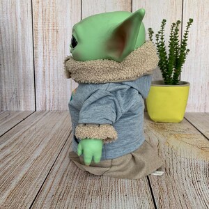 May include: A green-skinned doll with large ears, wearing a light blue hooded sweatshirt with tan trim and a tan skirt. The doll is posed in profile, with a small plant in a yellow pot in the background.