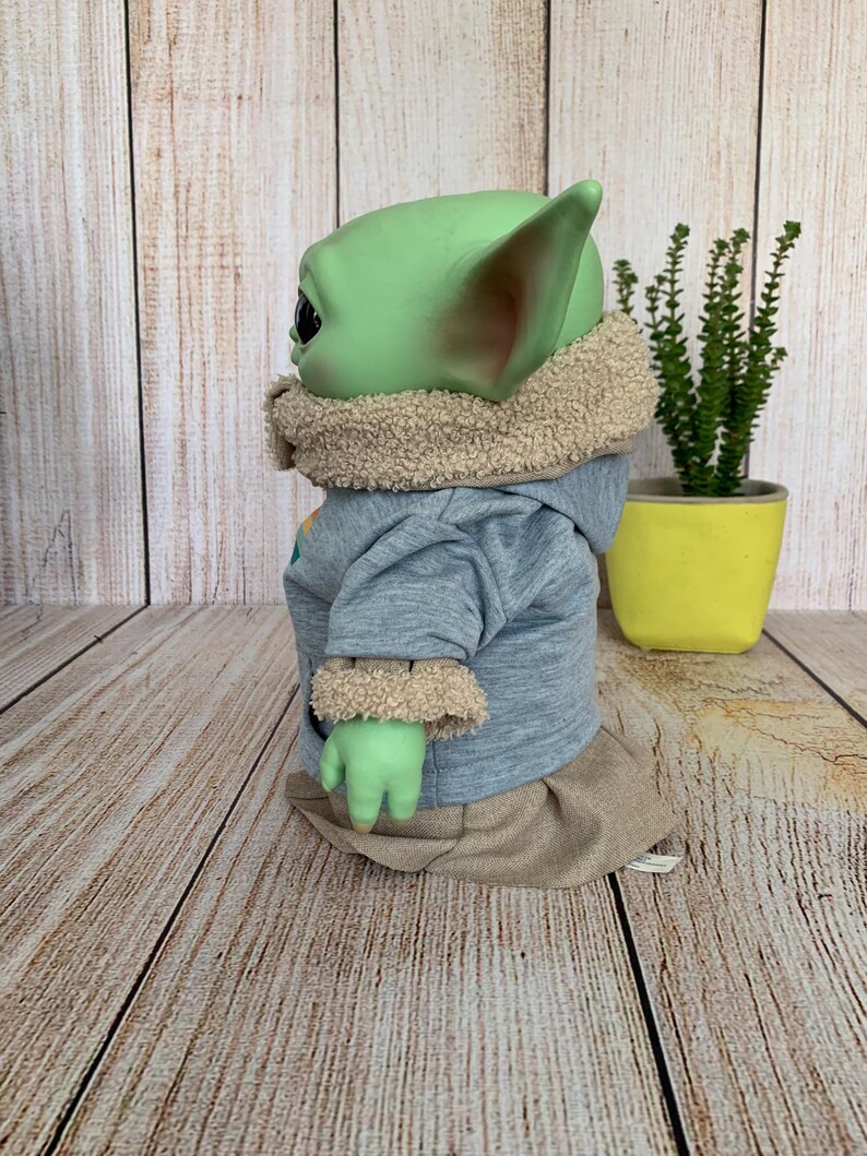 May include: A green-skinned doll with large ears, wearing a light blue hooded sweatshirt with tan trim and a tan skirt. The doll is posed in profile, with a small plant in a yellow pot in the background.