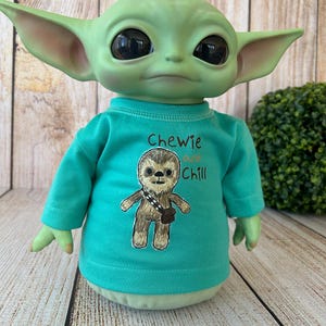 Peut inclure: Poup&eacute;e verte avec de grandes oreilles et de grands yeux, v&ecirc;tue d'un t-shirt turquoise. Le t-shirt pr&eacute;sente un dessin d'un Wookiee et le texte "Chewie and Chill". La poup&eacute;e est pos&eacute;e sur un fond en bois.