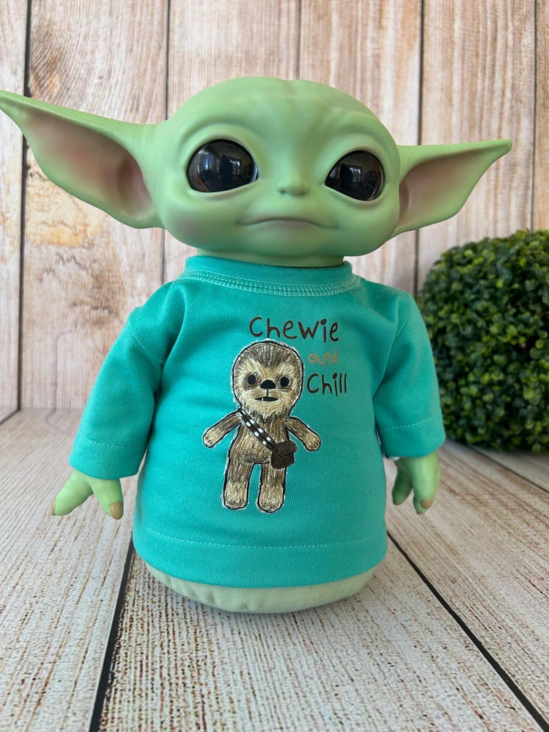 Peut inclure: Poup&eacute;e verte avec de grandes oreilles et de grands yeux, v&ecirc;tue d'un t-shirt turquoise. Le t-shirt pr&eacute;sente un dessin d'un Wookiee et le texte "Chewie and Chill". La poup&eacute;e est pos&eacute;e sur un fond en bois.