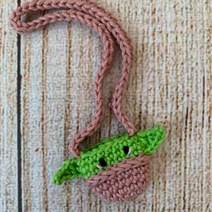 K&ouml;nnte beinhalten: Eine kleine geh&auml;kelte Tasche mit einem gr&uuml;nen Yoda-Charakterdesign. Die Tasche hat einen rosa Riemen und ist aus weichem Garn gefertigt.