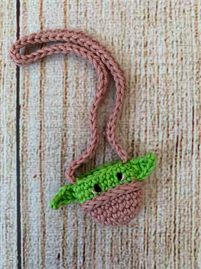 K&ouml;nnte beinhalten: Eine kleine geh&auml;kelte Tasche mit einem gr&uuml;nen Yoda-Charakterdesign. Die Tasche hat einen rosa Riemen und ist aus weichem Garn gefertigt.