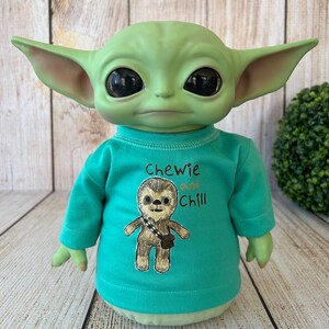 Peut inclure: Une poup&eacute;e verte ressemblant &agrave; un personnage d'une franchise de science-fiction populaire. La poup&eacute;e porte un t-shirt &agrave; manches longues turquoise avec les mots "Chewie and Chill" et un dessin d'une cr&eacute;ature ressemblant &agrave; un Wookiee. La poup&eacute;e a de grands yeux noirs et des oreilles pointues.
