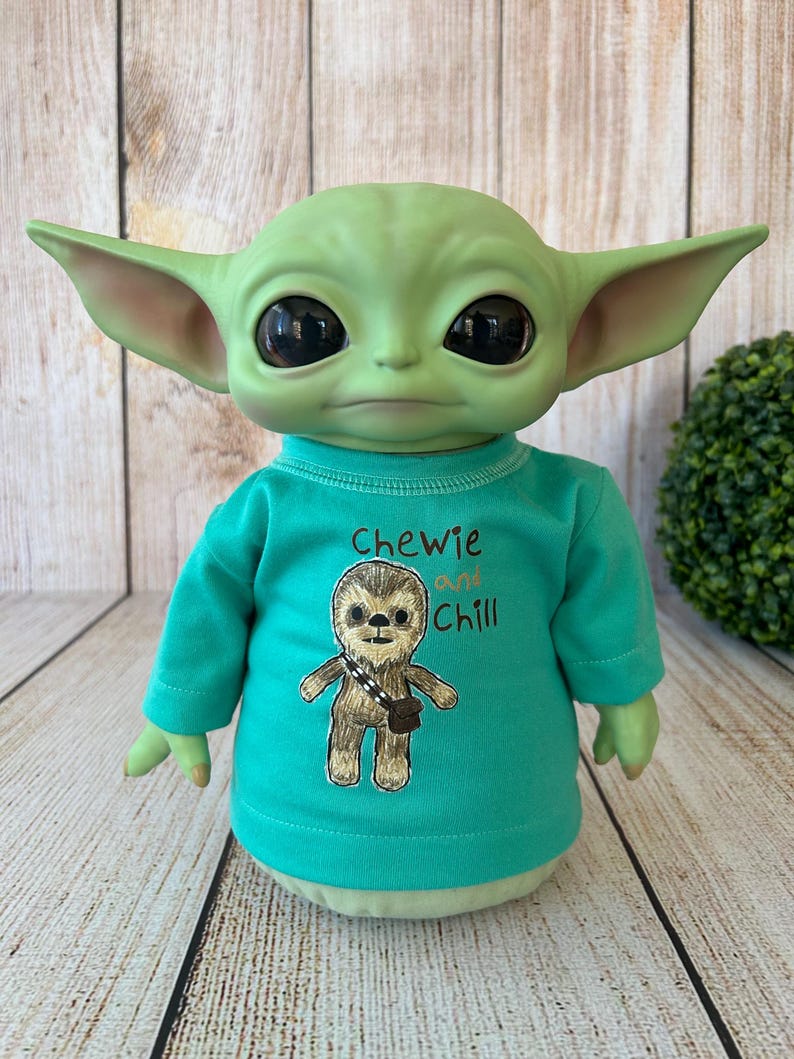 Peut inclure: Une poup&eacute;e verte ressemblant &agrave; un personnage d'une franchise de science-fiction populaire. La poup&eacute;e porte un t-shirt &agrave; manches longues turquoise avec les mots "Chewie and Chill" et un dessin d'une cr&eacute;ature ressemblant &agrave; un Wookiee. La poup&eacute;e a de grands yeux noirs et des oreilles pointues.