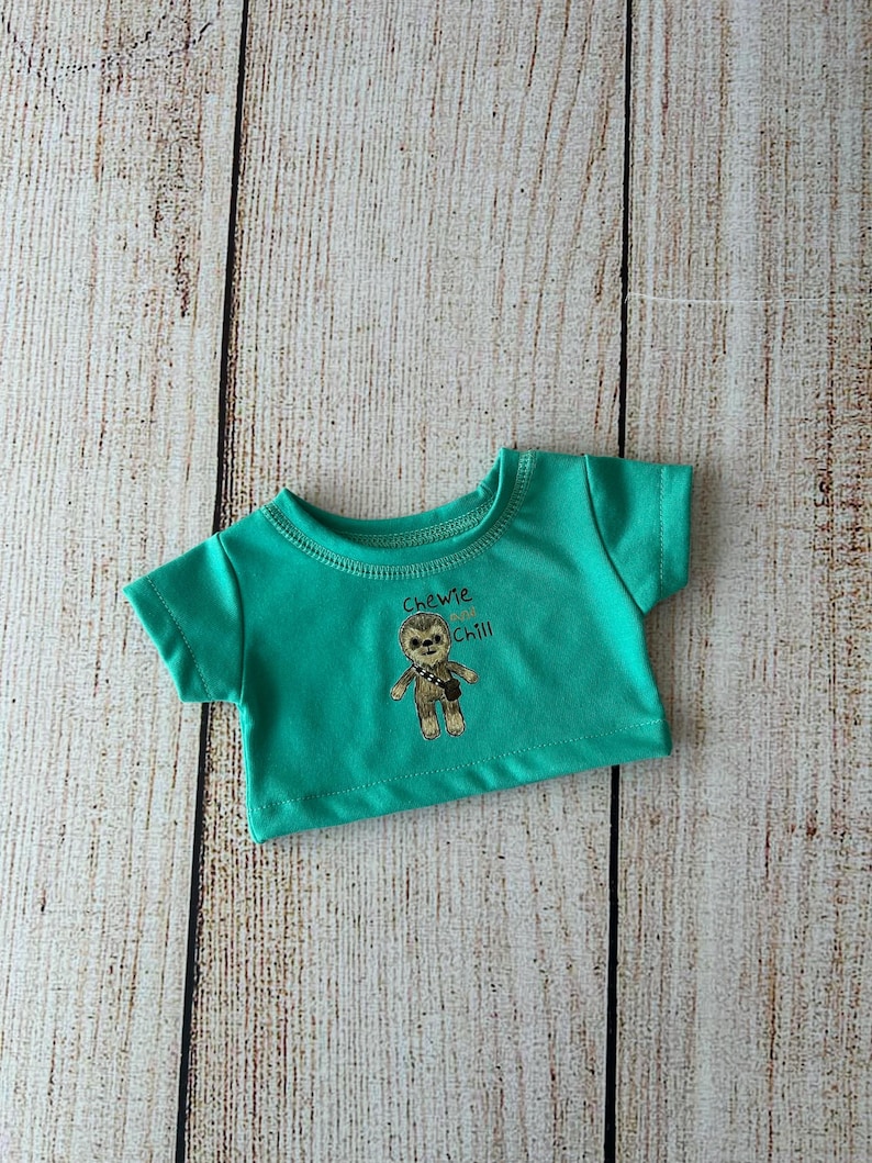 May include: A teal-colored t-shirt with a graphic of a cartoon character and the words "Chewie and Chill". The shirt is made of a soft material and has short sleeves. The shirt is designed for a doll.