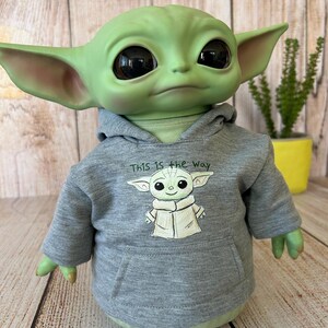 May include: A green figure with large black eyes and pointed ears, wearing a gray hoodie. The hoodie features a drawing of the figure and the text "This is the Way". The figure is a popular character from a science fiction series.