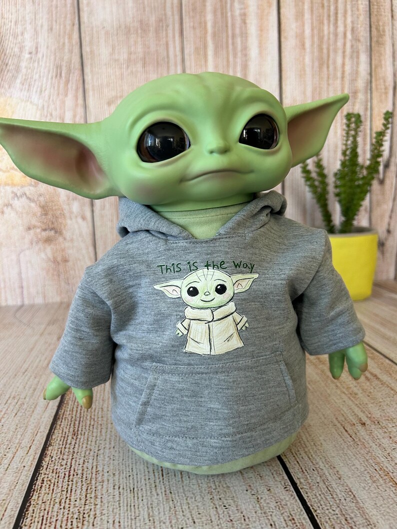 May include: A green figure with large black eyes and pointed ears, wearing a gray hoodie. The hoodie features a drawing of the figure and the text "This is the Way". The figure is a popular character from a science fiction series.