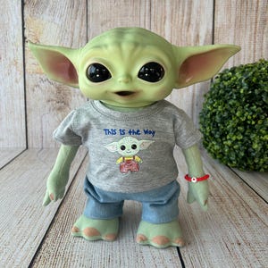 May include: A plush doll of a green alien character with large black eyes and pointed ears. The doll wears a gray t-shirt with the text "THIS IS THE WAY" and a cartoon image. It also has blue pants and a red bracelet.