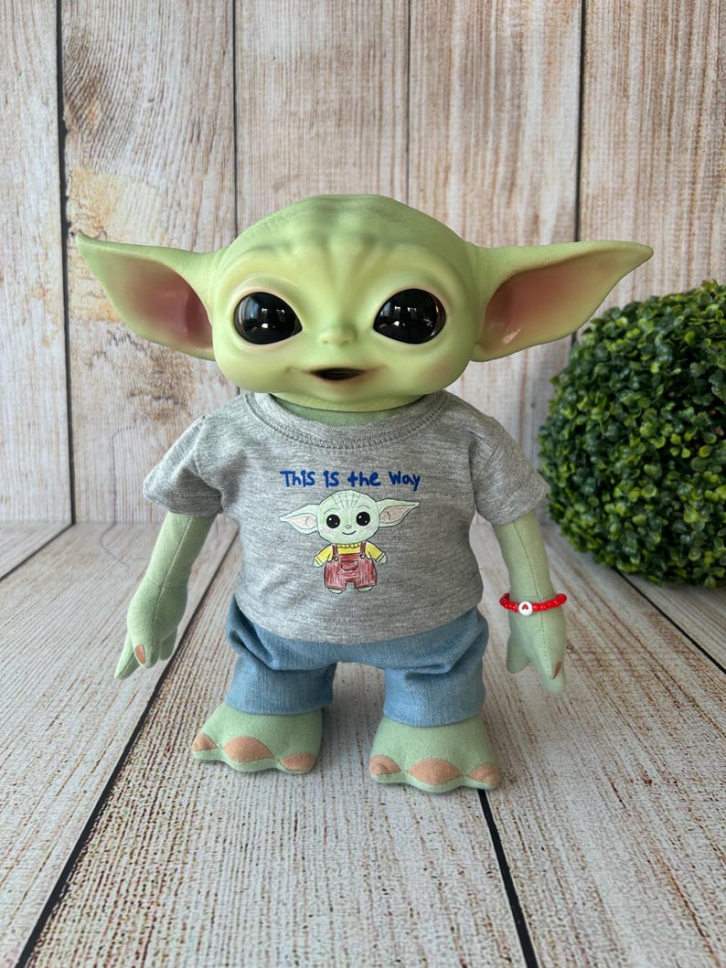 May include: A plush doll of a green alien character with large black eyes and pointed ears. The doll wears a gray t-shirt with the text "THIS IS THE WAY" and a cartoon image. It also has blue pants and a red bracelet.