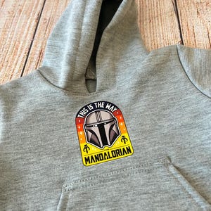 May include: A gray hooded sweatshirt featuring a Mandalorian graphic. The graphic includes the text "THIS IS THE WAY" and "THE MANDALORIAN" with a helmet design. The sweatshirt has a front pocket and a hood.