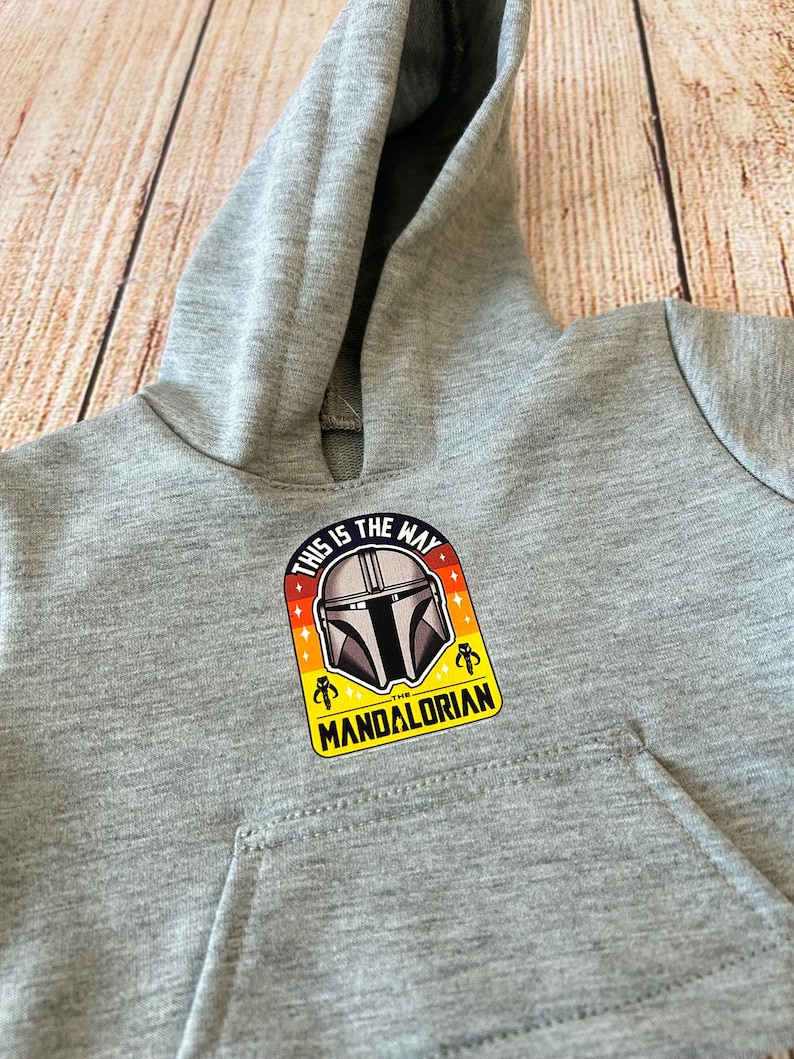 May include: A gray hooded sweatshirt featuring a Mandalorian graphic. The graphic includes the text "THIS IS THE WAY" and "THE MANDALORIAN" with a helmet design. The sweatshirt has a front pocket and a hood.