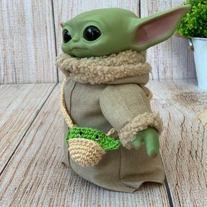 K&ouml;nnte beinhalten: Ein gr&uuml;nes Pl&uuml;schtier mit gro&szlig;en Augen, das einen braunen Mantel mit einem Kunstfellkragen und eine gr&uuml;n-beige geh&auml;kelte Tasche tr&auml;gt.