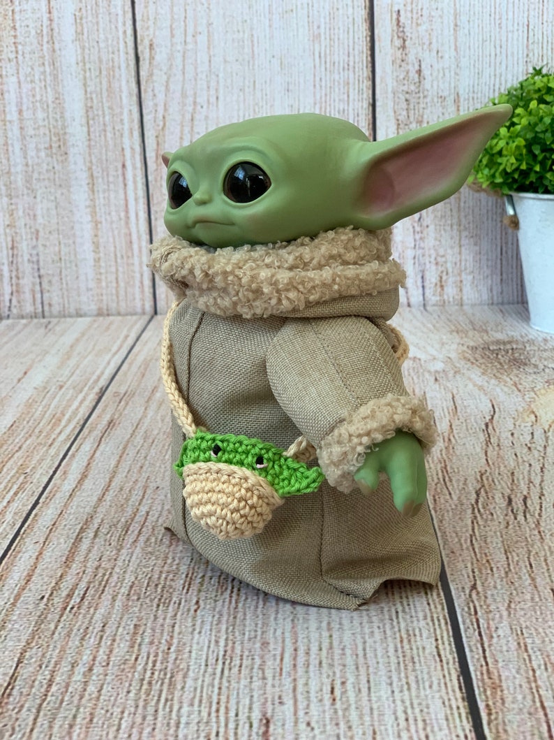 K&ouml;nnte beinhalten: Ein gr&uuml;nes Pl&uuml;schtier mit gro&szlig;en Augen, das einen braunen Mantel mit einem Kunstfellkragen und eine gr&uuml;n-beige geh&auml;kelte Tasche tr&auml;gt.