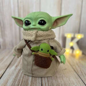 May include: A green plush toy with large eyes wearing a brown coat and a brown crocheted bag with a green plush toy inside.