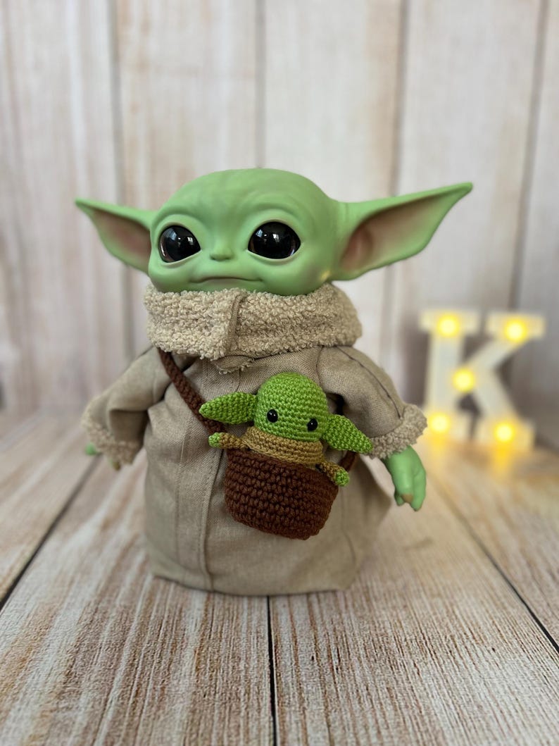 May include: A green plush toy with large eyes wearing a brown coat and a brown crocheted bag with a green plush toy inside.