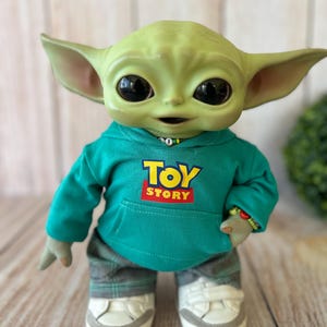 Op de afbeelding: Een groen wezenfiguurtje met grote zwarte ogen en puntige oren, gekleed in een blauwgroene hoodie met de tekst TOY STORY in geel en rood op de voorkant, een grijze broek met ruitjespatroon en witte sneakers met grijze accenten op de zolen.