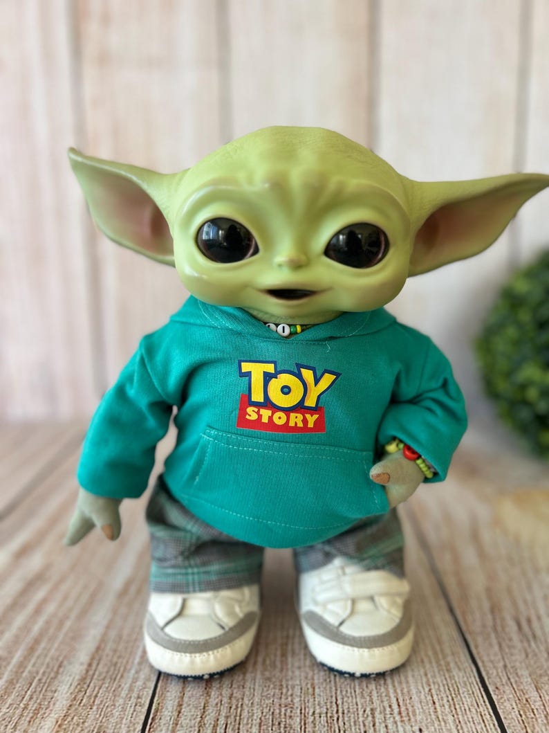 Op de afbeelding: Een groen wezenfiguurtje met grote zwarte ogen en puntige oren, gekleed in een blauwgroene hoodie met de tekst TOY STORY in geel en rood op de voorkant, een grijze broek met ruitjespatroon en witte sneakers met grijze accenten op de zolen.