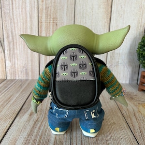 May include: A green-skinned creature figure with large ears wearing a striped knit sweater, blue denim pants with white stitching, and a small black backpack featuring a repeating pattern of grey helmets and green creature faces on the back.